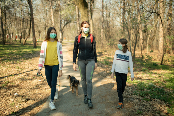 Mother with her children walking in public park with their mixed breed dog in time of corona virus outbreak. They wear protective mask and keeping social distance with other people as a prevention for spreading disease.