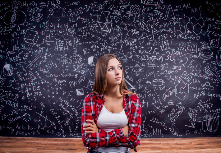 Beautiful student in red checked shirt against big blackboard with mathematical symbols and formulas. Studio shot on black background.