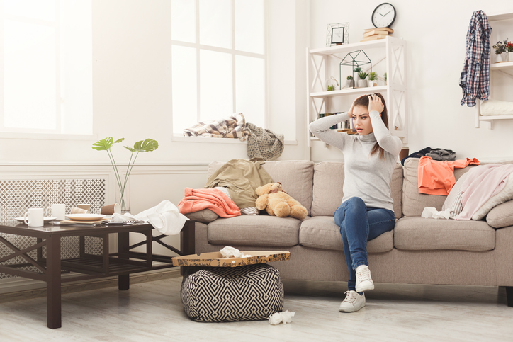 Desperate helpless woman sitting on sofa in messy living room. Young girl surrounded by many stack of clothes. Disorder and mess at home, copy space